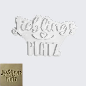 Op de afbeelding: Een wit, uitgesneden bord met de Duitse woorden "Lieblings Platz" in een scriptlettertype. Het woord "Lieblings" is boven "Platz" gebogen, met een hartsymbool ertussen. Een kleinere, vierkante afbeelding van de stempel staat in de linker benedenhoek.