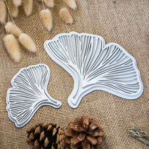 Può includere: Due stencil di foglie di ginkgo in metallo bianco, uno più grande dell'altro, su uno sfondo di juta marrone.