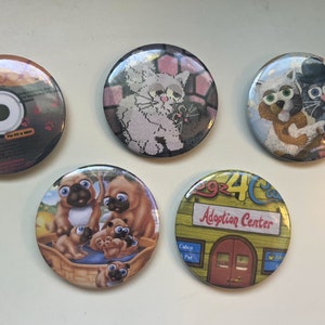 Op de afbeelding: Vijf ronde knopen met verschillende dierenontwerpen. De knopen tonen een zwart-witte kat, een zwart-witte kat met een hoge hoed, een groep mopshonden, een witte kat met een zwarte kat en een geel gebouw met de tekst "2924C Adoption Center".