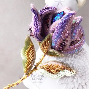 May include: A purple and pink beaded flower brooch with gold accents and green leaves. The brooch is on a white surface with a floral pattern. "OKSANA KONAK accessories" is written on the brooch.