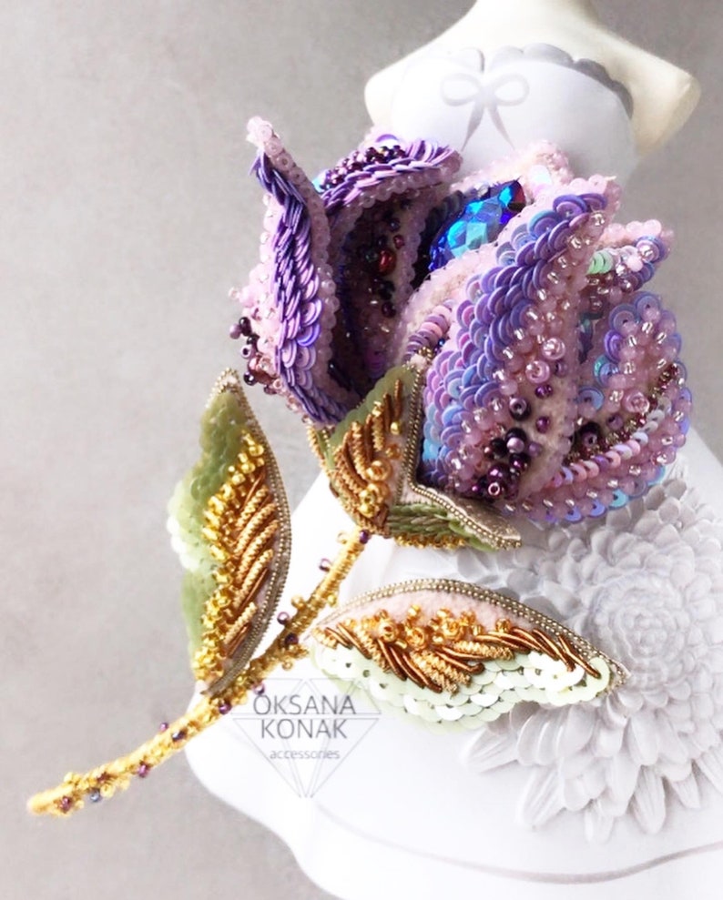 May include: A purple and pink beaded flower brooch with gold accents and green leaves. The brooch is on a white surface with a floral pattern. "OKSANA KONAK accessories" is written on the brooch.