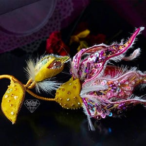 K&ouml;nnte beinhalten: Eine Brosche in Form einer rosa und gelben Blume mit Pailletten und Perlen. Die Blume ist aus Stoff gefertigt und hat ein zartes, feminines Design. Die Brosche ist mit "Oksana Konak" signiert.