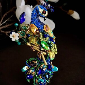 May include: A peacock brooch made of blue, green, and gold beads and sequins. The peacock is perched on a branch with a flower and leaves. The brooch is a beautiful and intricate piece of jewelry.