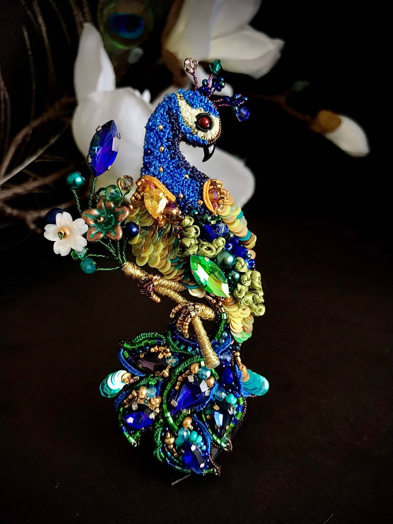 May include: A peacock brooch made of blue, green, and gold beads and sequins. The peacock is perched on a branch with a flower and leaves. The brooch is a beautiful and intricate piece of jewelry.
