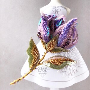 May include: A delicate, hand-beaded rose brooch with iridescent purple and pink sequins, gold accents, and green leaves. The brooch is displayed on a white mannequin.
