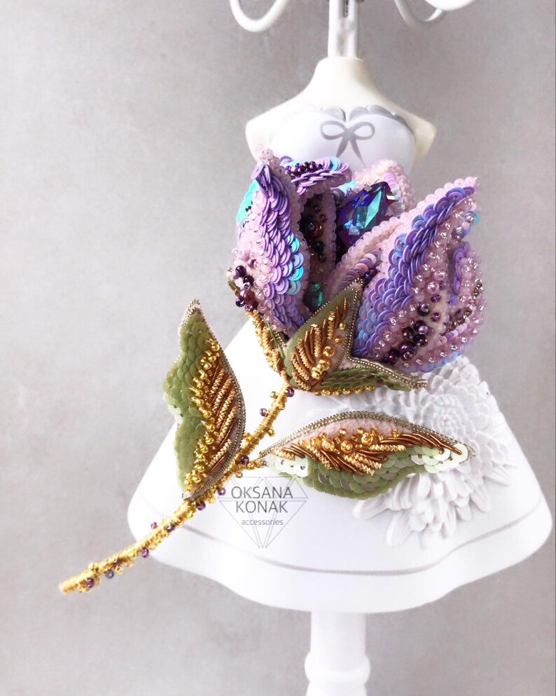 May include: A delicate, hand-beaded rose brooch with iridescent purple and pink sequins, gold accents, and green leaves. The brooch is displayed on a white mannequin.