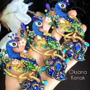 May include: Three peacock brooches made of blue, green, and gold beads. The brooches are on a hand and feature a floral design. The text "Oksana Konak" is visible in the bottom right corner of the image.