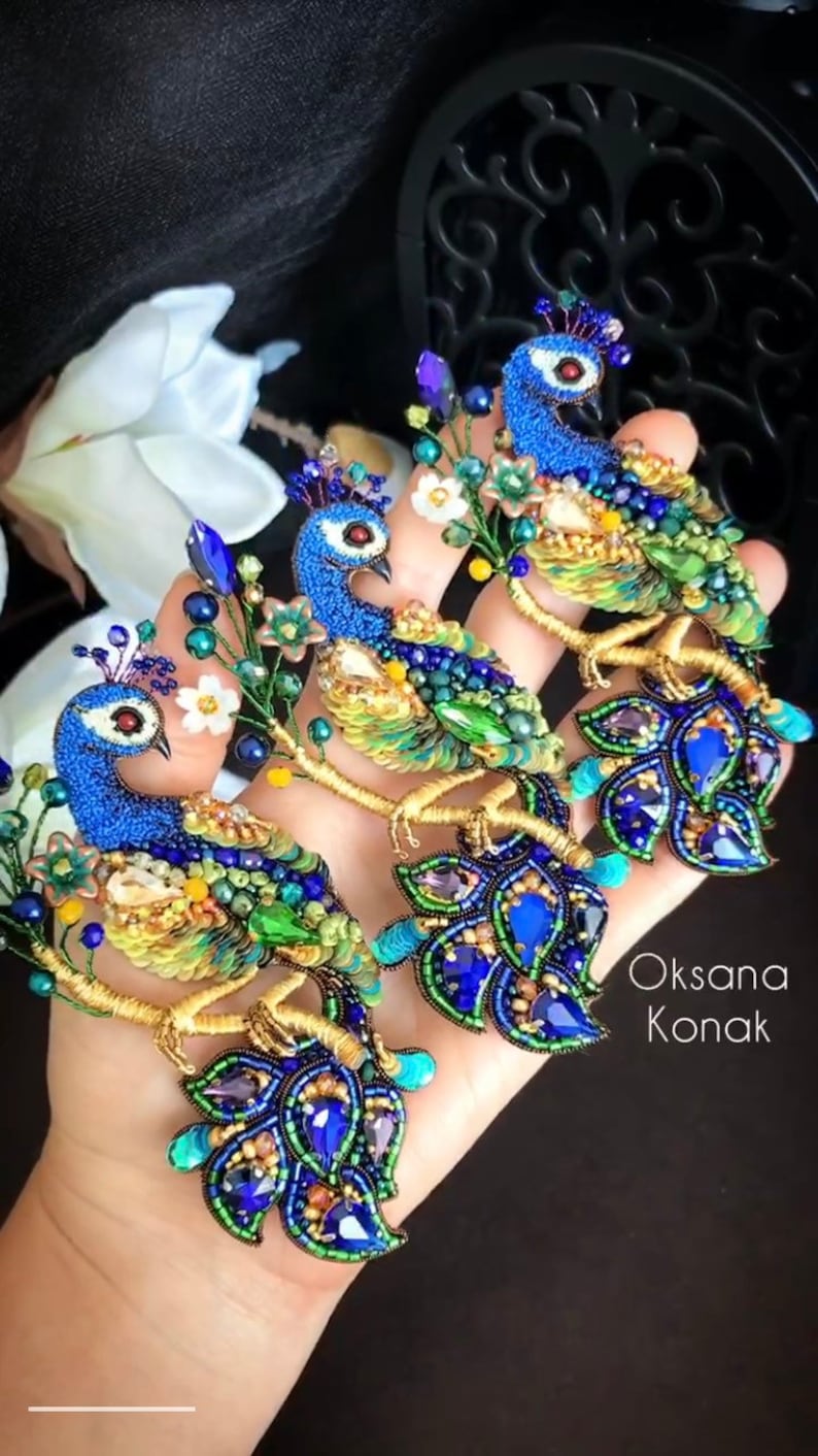 May include: Three peacock brooches made of blue, green, and gold beads. The brooches are on a hand and feature a floral design. The text "Oksana Konak" is visible in the bottom right corner of the image.