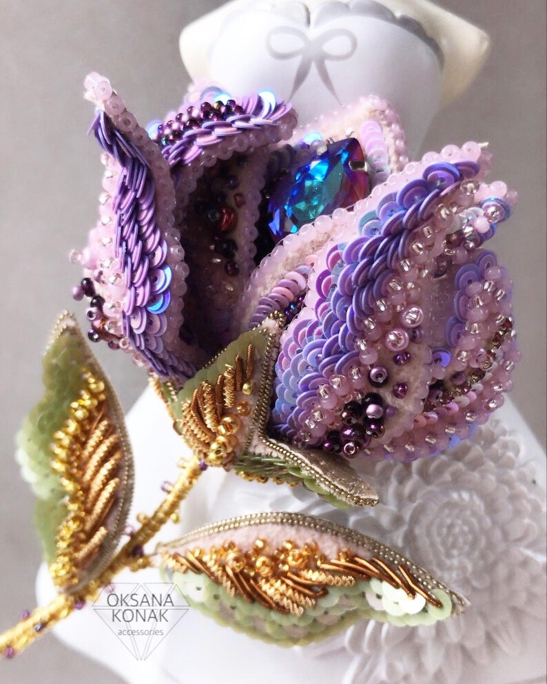 May include: A delicate purple and pink rose brooch with a sparkling blue crystal centre, embellished with sequins and beads. The rose is attached to a gold and green stem with leaves.
