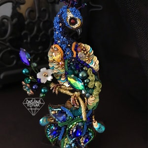 May include: A beaded peacock brooch with blue, green, gold, and white accents. The peacock is perched on a leafy branch with a crown on its head. The brooch is made by Oksana Konak accessories.