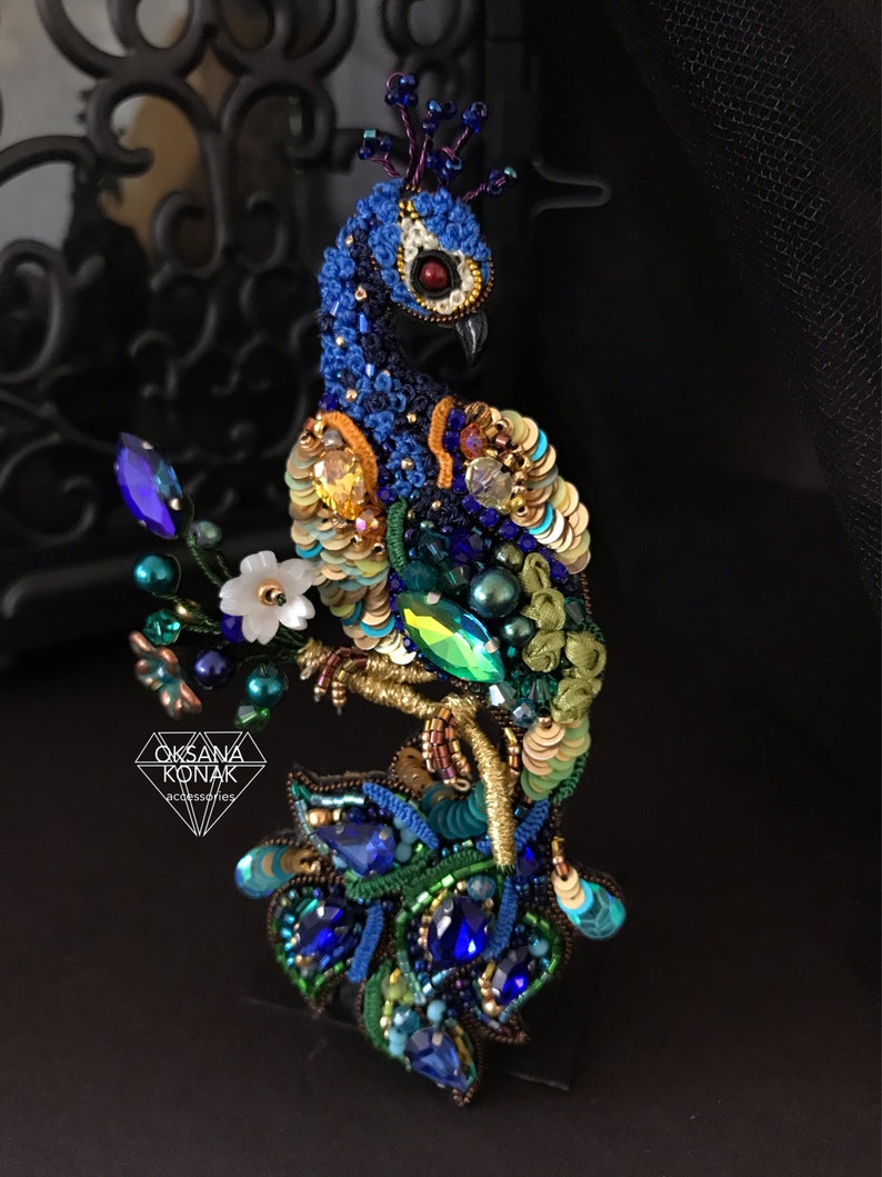 May include: A beaded peacock brooch with blue, green, gold, and white accents. The peacock is perched on a leafy branch with a crown on its head. The brooch is made by Oksana Konak accessories.
