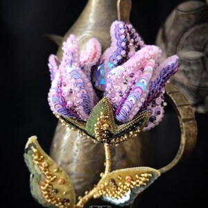 May include: A delicate pink and purple flower brooch with iridescent sequins and beads. The flower is beaded with a green stem and leaves. The brooch is handmade and features the text "OKSANA KONAK accessories".