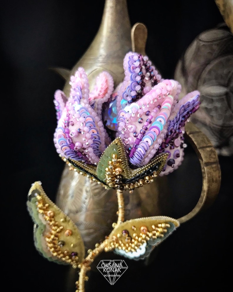May include: A delicate pink and purple flower brooch with iridescent sequins and beads. The flower is beaded with a green stem and leaves. The brooch is handmade and features the text "OKSANA KONAK accessories".
