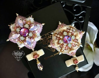 May include: A pair of pink and cream beaded earrings with a floral design. The earrings feature a large, iridescent crystal in the center of each flower. The earrings are accented with small pearls and gold beads. The earrings are hanging from a small bow.