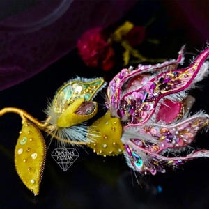 K&ouml;nnte beinhalten: Eine Brosche in Form einer rosa und gelben Blume mit wei&szlig;em Fell und goldenen Akzenten. Die Brosche ist mit Pailletten und Perlen verziert.