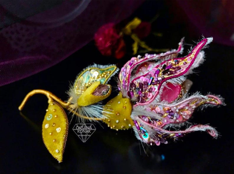 K&ouml;nnte beinhalten: Eine Brosche in Form einer rosa und gelben Blume mit wei&szlig;em Fell und goldenen Akzenten. Die Brosche ist mit Pailletten und Perlen verziert.