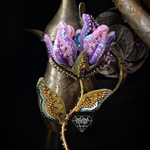 May include: A beaded flower brooch featuring a purple, pink, and green flower with gold accents. The brooch is displayed against a gold metal background. The text "OKSANA KONAK accessories" is at the bottom of the image.