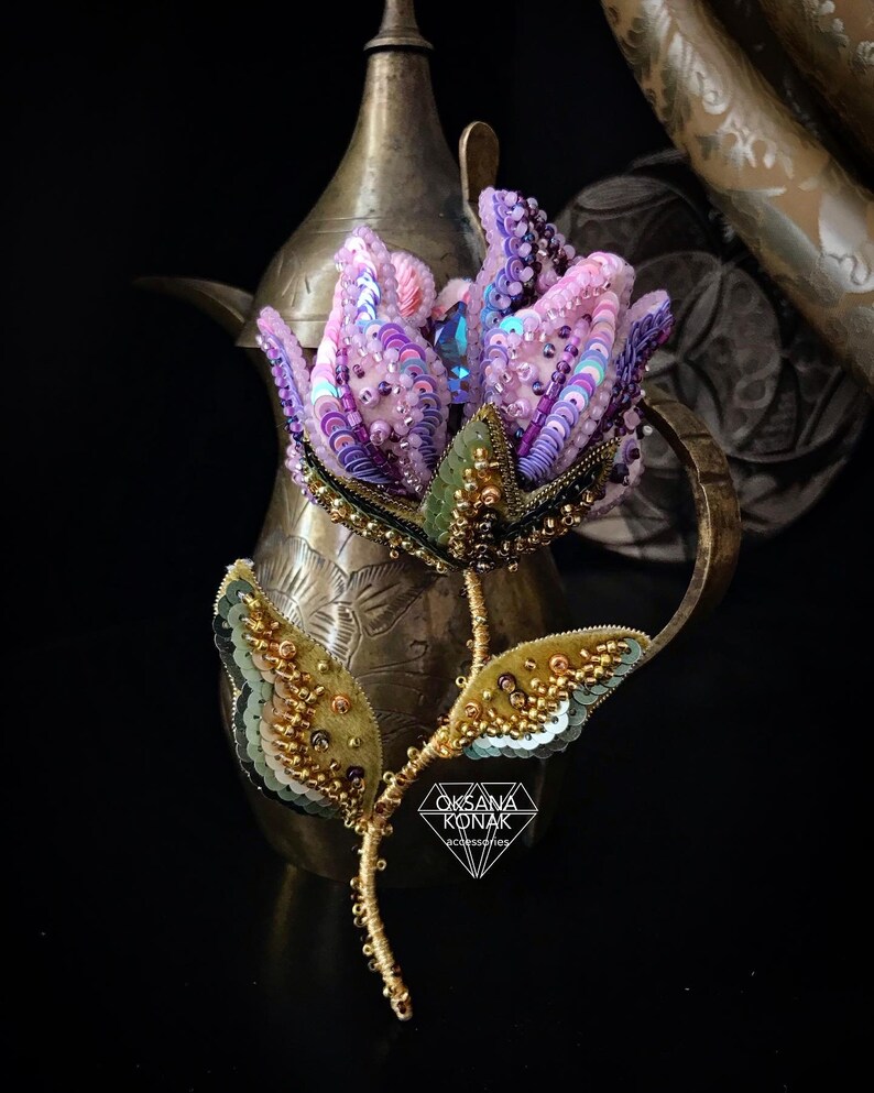 May include: A beaded flower brooch featuring a purple, pink, and green flower with gold accents. The brooch is displayed against a gold metal background. The text "OKSANA KONAK accessories" is at the bottom of the image.