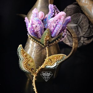 May include: A delicate, hand-beaded flower brooch featuring a pink and purple flower, green leaves, and gold accents. The brooch is displayed on a vintage brass teapot.