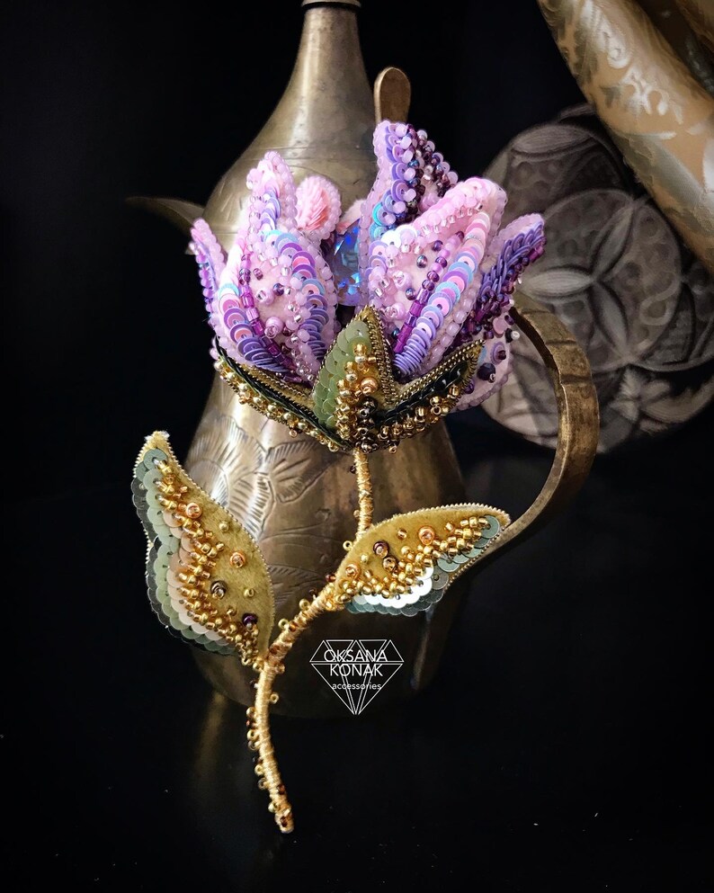 May include: A delicate, hand-beaded flower brooch featuring a pink and purple flower, green leaves, and gold accents. The brooch is displayed on a vintage brass teapot.