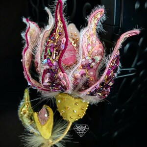 K&ouml;nnte beinhalten: Eine Brosche in Form einer rosa und violetten Blume mit goldenen und wei&szlig;en Akzenten. Die Blume besteht aus Stoff und Perlen und hat eine flauschige Textur. Die Brosche ist an einem goldenen Stiel mit einem gr&uuml;nen Blatt befestigt.