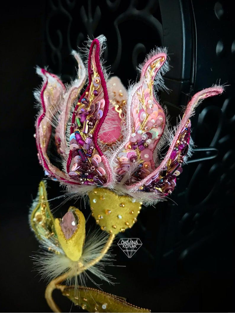 K&ouml;nnte beinhalten: Eine Brosche in Form einer rosa und violetten Blume mit goldenen und wei&szlig;en Akzenten. Die Blume besteht aus Stoff und Perlen und hat eine flauschige Textur. Die Brosche ist an einem goldenen Stiel mit einem gr&uuml;nen Blatt befestigt.