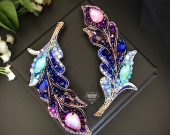 May include: A pair of beaded feather brooches in shades of blue, purple, and pink. The brooches are displayed on a black velvet background.