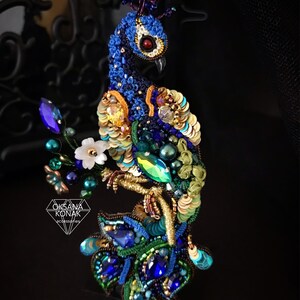 May include: A blue and green peacock brooch with a crown, made with sequins, beads, and embroidery. The peacock is perched on a branch with white flowers.