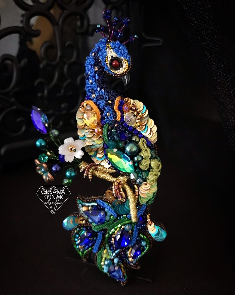 May include: A blue and green peacock brooch with a crown, made with sequins, beads, and embroidery. The peacock is perched on a branch with white flowers.
