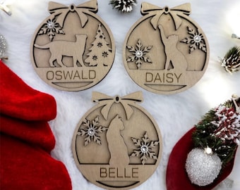 May include: Three wooden Christmas ornaments shaped like a ball with a cat silhouette, snowflakes, and a bow. Each ornament has a different cat breed and name: Oswald, Daisy, and Belle.
