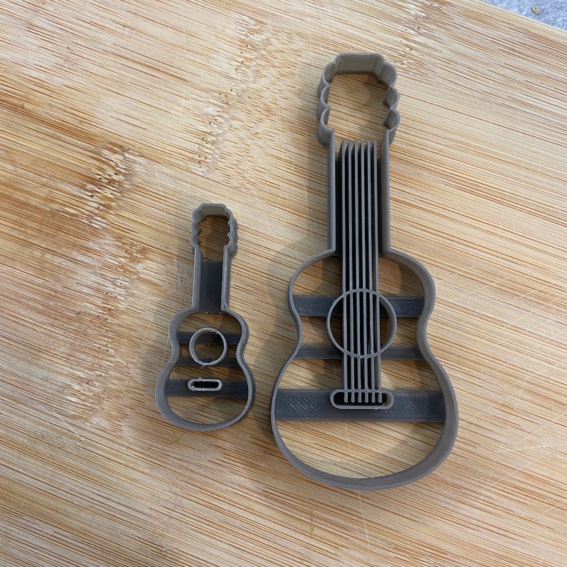 Puede incluir: Dos cortadores de galletas de guitarra de metal gris, uno grande y otro peque&ntilde;o. El cortador m&aacute;s grande tiene un dise&ntilde;o detallado con cuerdas y un agujero de sonido. El cortador m&aacute;s peque&ntilde;o es un dise&ntilde;o simplificado.