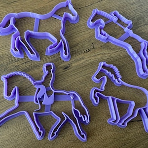 May include: Four purple plastic horse cookie cutters. The cutters are in the shape of horses, one with a rider, and one with a raised leg.