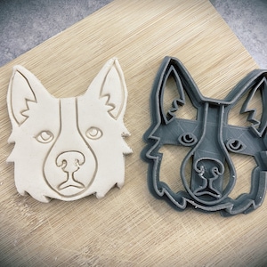 May include: Two dog-shaped cookie cutters, one white and one grey, on a wooden surface. The cutters are detailed with ears, eyes, and a nose.
