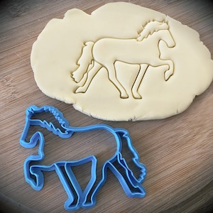 May include: A blue plastic cookie cutter shaped like a horse. The cutter is on a wooden surface next to a piece of dough that has been cut out with the cutter.
