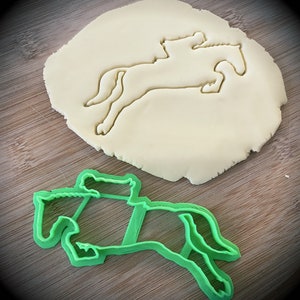 May include: Green plastic cookie cutter shaped like a horse jumping over a hurdle. The cutter is on a wooden surface with a white dough cutout of the same design.