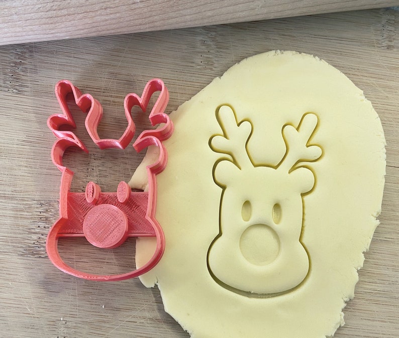 May include: A red plastic reindeer cookie cutter with a white cookie dough cutout of the same design.