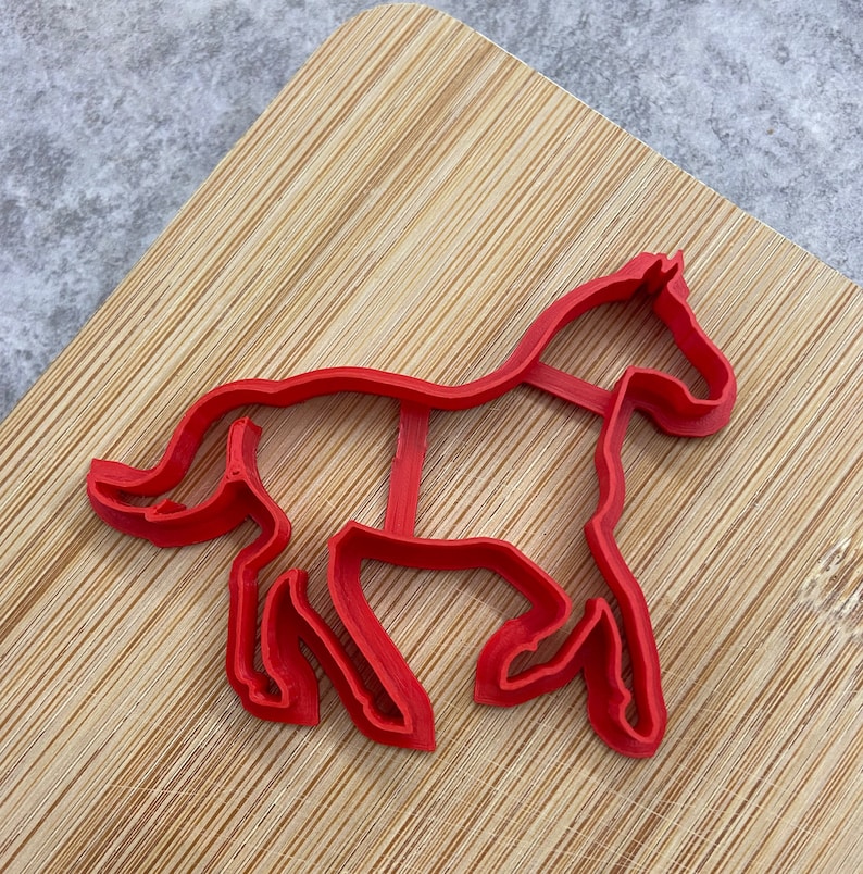 Pu&ograve; includere: Tagliapasta a forma di cavallo in plastica rossa su una superficie di legno.