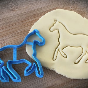 Pu&ograve; includere: Un tagliapasta a forma di cavallo in plastica blu su una superficie di legno, accanto a un pezzo di pasta per biscotti con la forma del cavallo tagliata.