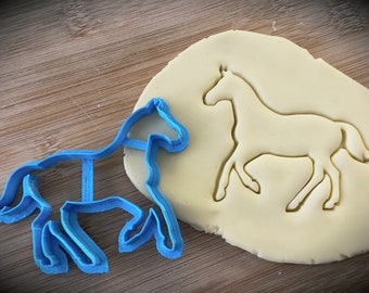 May include: A blue plastic horse cookie cutter on a wooden surface, next to a piece of cookie dough with the horse shape cut out.