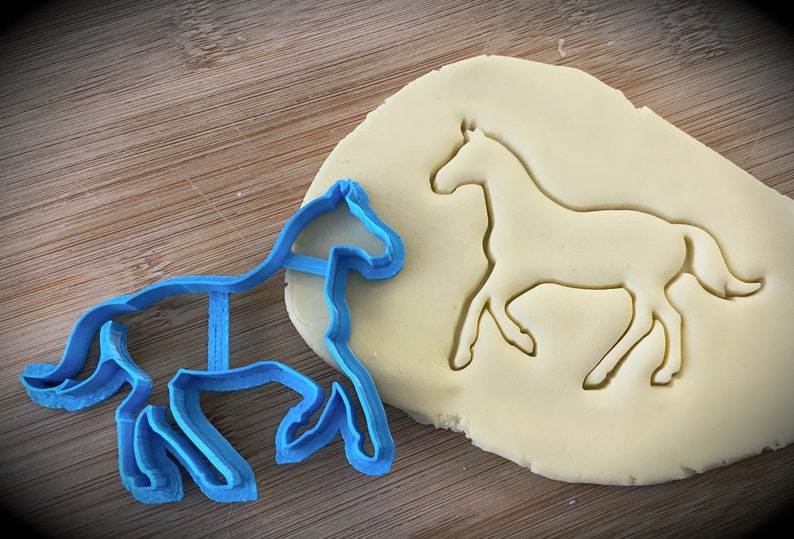 Pu&ograve; includere: Un tagliapasta a forma di cavallo in plastica blu su una superficie di legno, accanto a un pezzo di pasta per biscotti con la forma del cavallo tagliata.