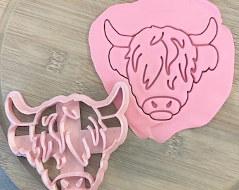 May include: Two pink plastic cookie cutters shaped like Highland cows. The cutters are on a wooden surface.