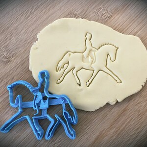 May include: A blue plastic cookie cutter in the shape of a person riding a horse. The cutter is on a wooden surface next to a piece of dough with the same shape cut out.