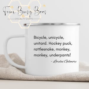 Puede incluir: Taza de esmalte blanco con borde y asa negros. La taza presenta el texto "Bicycle, unicycle, unitard. Hockey puck, rattlesnake, monkey, monkey, underpants!" atribuido a Lorelai Gilmore. La taza tiene el logo "Four Busy Bees".
