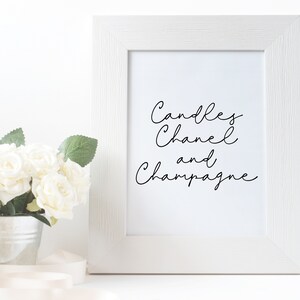 以下が含まれることがあります： 黒い筆記体で「Candles Chanel and Champagne」と書かれた白い額縁入りプリント