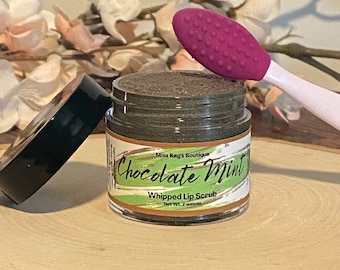 Może przedstawiać: Słoik "Chocolate Mint" peelingu do ust z różową szczoteczką do peelingu ust na drewnianym plastrze. Słoik ma czarną pokrywkę i etykietę z zielonymi akcentami. Tło obejmuje białe i różowe kwiaty.