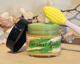 Może przedstawiać: Słoik zielonego peelingu do ust z napisem "Caramel Apple" i "Whipped Lip Scrub" na etykiecie. Słoik jest otwarty, z czarną pokrywką obok. Żółto-różowy aplikator do peelingu do ust znajduje się obok słoika. Słoik stoi na drewnianej powierzchni.