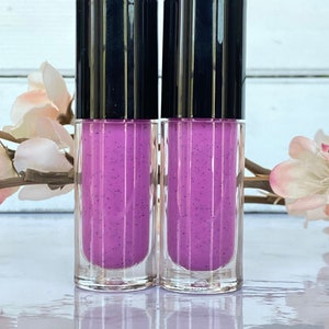 May include: Two clear glass tubes with black caps, filled with a vibrant purple liquid speckled with dark particles. The tubes are displayed on a reflective surface, with soft pink flowers in the background.