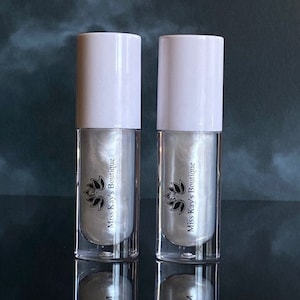 Könnte beinhalten: Zwei klare, zylindrische Flaschen mit flüssigem Schimmer und weißen Kappen. Die Flaschen haben einen silbernen Schimmer im Inneren und ein Logo mit der Aufschrift "Miss Kay's Boutique". Die Flaschen spiegeln sich auf einer dunklen Oberfläche.