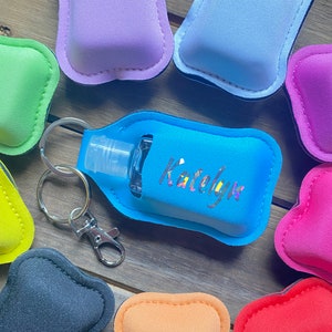 Könnte beinhalten: Eine Sammlung farbenfroher Handdesinfektionsmittelhalter mit Schlüsselanhängern. Ein hellblauer Halter trägt den Namen "Katelyn" in silbernen Buchstaben. Weitere Halter sind in Pink-, Lila-, Grün-, Orange- und Grautönen gehalten. Die Halter sind aus weichem, gepolstertem Material.