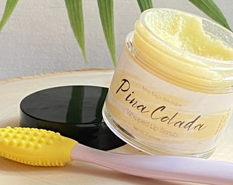 Może przedstawiać: Słoik z peelingiem do ust Pina Colada z żółtym aplikatorem. Słoik ma czarną pokrywkę i etykietę z nazwą produktu i marką. Peeling do ust ma jasnożółty kolor i waży 57 gramów.